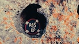 A round object resembling a lifebuoy is placed inside a rough, rocky cavity. The object is dark with red and white accents and features white painted letters 'ReS KP' on its surface. The surrounding rock has a textured appearance, with patches of orange and grey tones.