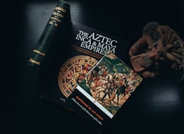 An assortment of books and artifacts related to Mesoamerican history are arranged on a dark surface. Notably, a book titled 'The Aztec, Inca & Maya Empires' featuring a vibrant Aztec calendar on the cover, alongside another book depicting a historical painting of Spanish conquest. A small clay sculpture resembling an ancient Mesoamerican figure is positioned nearby.