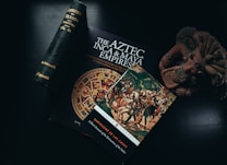 An assortment of books and artifacts related to Mesoamerican history are arranged on a dark surface. Notably, a book titled 'The Aztec, Inca & Maya Empires' featuring a vibrant Aztec calendar on the cover, alongside another book depicting a historical painting of Spanish conquest. A small clay sculpture resembling an ancient Mesoamerican figure is positioned nearby.