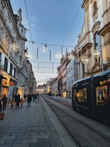 A European city street with historic architecture, illuminated by decorative lights hanging across the street. A tram is visible on the right, and people walk along the sidewalks, engaging in shopping and strolling. The sky is clear, hinting at a calm evening atmosphere.