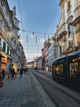 A European city street with historic architecture, illuminated by decorative lights hanging across the street. A tram is visible on the right, and people walk along the sidewalks, engaging in shopping and strolling. The sky is clear, hinting at a calm evening atmosphere.