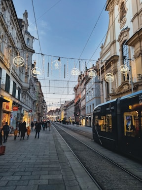 A European city street with historic architecture, illuminated by decorative lights hanging across the street. A tram is visible on the right, and people walk along the sidewalks, engaging in shopping and strolling. The sky is clear, hinting at a calm evening atmosphere.