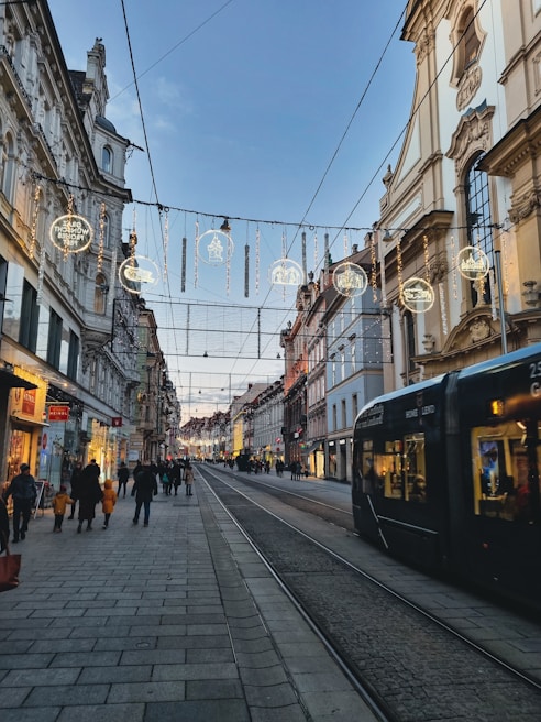 A European city street with historic architecture, illuminated by decorative lights hanging across the street. A tram is visible on the right, and people walk along the sidewalks, engaging in shopping and strolling. The sky is clear, hinting at a calm evening atmosphere.