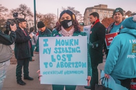 A group of people participating in a protest or demonstration, with a woman in the foreground holding a sign that reads 'I Mourn My Sibling Lost to Abortion.' Other people around are also holding signs and wearing clothing with messages related to the protest. A cameraman is filming the scene, and there is a building visible in the background.
