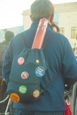 A person facing away, wearing a backpack decorated with several activist buttons, including messages about climate change, feminism, and political criticism. A rolled-up poster is tucked into the top of the backpack. The setting appears to be outdoors with other people in the background.