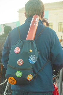 A person facing away, wearing a backpack decorated with several activist buttons, including messages about climate change, feminism, and political criticism. A rolled-up poster is tucked into the top of the backpack. The setting appears to be outdoors with other people in the background.