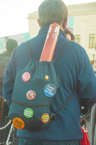 A person facing away, wearing a backpack decorated with several activist buttons, including messages about climate change, feminism, and political criticism. A rolled-up poster is tucked into the top of the backpack. The setting appears to be outdoors with other people in the background.