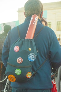 A person facing away, wearing a backpack decorated with several activist buttons, including messages about climate change, feminism, and political criticism. A rolled-up poster is tucked into the top of the backpack. The setting appears to be outdoors with other people in the background.