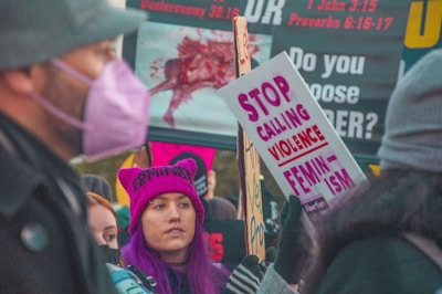 A group of people participating in a protest, holding signs with messages about feminism and violence. One person with purple hair and a matching hat is prominently featured, while another person wears a mask. The background includes more signage with text.