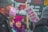 A group of people participating in a protest, holding signs with messages about feminism and violence. One person with purple hair and a matching hat is prominently featured, while another person wears a mask. The background includes more signage with text.