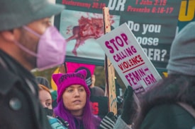 A group of people participating in a protest, holding signs with messages about feminism and violence. One person with purple hair and a matching hat is prominently featured, while another person wears a mask. The background includes more signage with text.