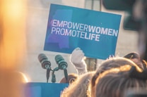 A close-up of a crowd with someone holding a blue sign with white text that reads 'EMPOWER WOMEN PROMOTE LIFE'. The sign is brightly lit by sunlight from the left, and multiple microphones are visible in the foreground.