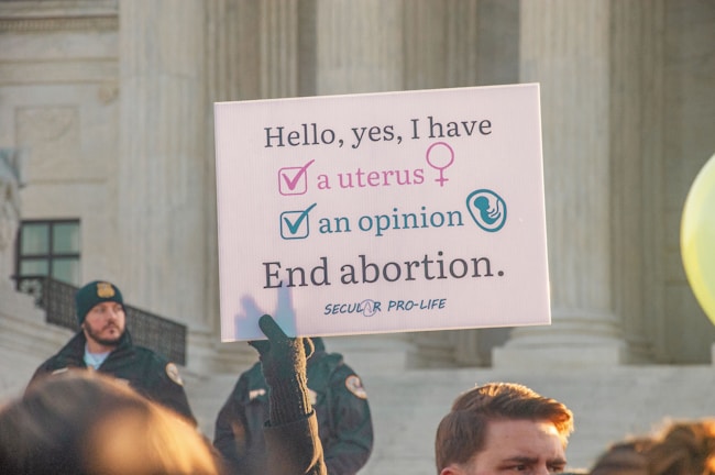 A protest sign held by someone in front of a large stone building with columns, advocating a pro-life message related to abortion. The sign reads, 'Hello, yes, I have a uterus, an opinion. End abortion.' A crowd is gathered, and some individuals are visible wearing security or official uniforms.