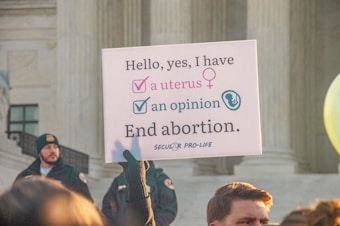 A protest sign held by someone in front of a large stone building with columns, advocating a pro-life message related to abortion. The sign reads, 'Hello, yes, I have a uterus, an opinion. End abortion.' A crowd is gathered, and some individuals are visible wearing security or official uniforms.