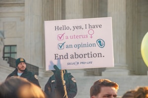 A protest sign held by someone in front of a large stone building with columns, advocating a pro-life message related to abortion. The sign reads, 'Hello, yes, I have a uterus, an opinion. End abortion.' A crowd is gathered, and some individuals are visible wearing security or official uniforms.