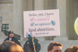 A protest sign held by someone in front of a large stone building with columns, advocating a pro-life message related to abortion. The sign reads, 'Hello, yes, I have a uterus, an opinion. End abortion.' A crowd is gathered, and some individuals are visible wearing security or official uniforms.