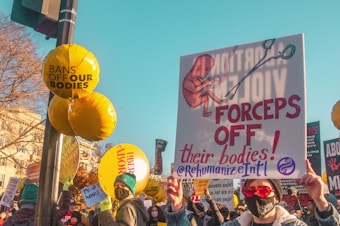 A group of protesters holding various signs advocating for reproductive rights. Prominent signs include one with a fetus and the text 'FORCEPS OFF their bodies!' and another that reads 'BANS OFF OUR BODIES'. People in the crowd are wearing masks and hats, with a clear blue sky in the background.