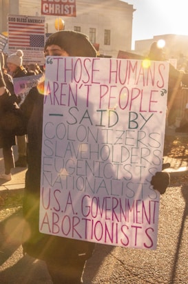 A person is holding a protest sign with bold, colorful letters in front of a small crowd. The sign criticizes various groups like colonizers and nationalists for dehumanizing others. Other protesters and signs are visible, conveying a sense of activism.