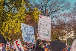 A group of people hold up protest signs in an outdoor setting. The signs contain messages advocating life and opposing killing by various authorities. Trees with autumn foliage can be seen in the background.
