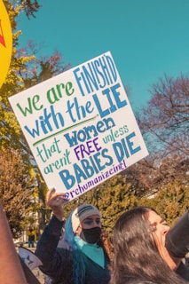 A person in a black mask and beanie holds a protest sign with colorful text in a public park-like setting. The sign contains a message about women's rights. Another person beside them appears to be speaking or shouting into a microphone, indicating expressions of protest.