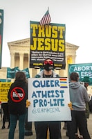 A protest scene outside a prominent building with classical architecture, featuring individuals holding various signs. One sign reads 'TRUST JESUS' with large, bold, yellow letters, topped with a small American flag. In front, a person holds a sign stating 'QUEER ATHEIST PRO-LIFE' with colorful stripes representing different flags.