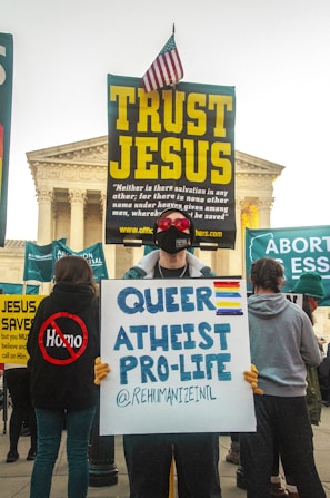 A protest scene outside a prominent building with classical architecture, featuring individuals holding various signs. One sign reads 'TRUST JESUS' with large, bold, yellow letters, topped with a small American flag. In front, a person holds a sign stating 'QUEER ATHEIST PRO-LIFE' with colorful stripes representing different flags.