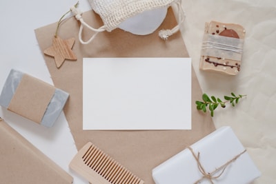 A flat lay composition featuring natural and eco-friendly items such as handmade soaps wrapped in brown paper, a wooden comb, a small plant branch, and a star-shaped wooden ornament. The items are arranged around a blank white card on a brown craft paper background, with a netted pouch visible at the top.