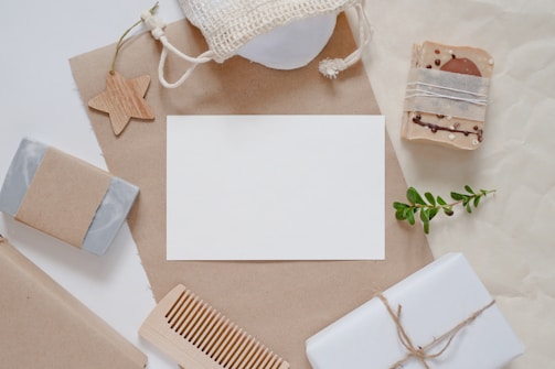 A flat lay composition featuring natural and eco-friendly items such as handmade soaps wrapped in brown paper, a wooden comb, a small plant branch, and a star-shaped wooden ornament. The items are arranged around a blank white card on a brown craft paper background, with a netted pouch visible at the top.