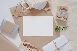 A flat lay composition featuring natural and eco-friendly items such as handmade soaps wrapped in brown paper, a wooden comb, a small plant branch, and a star-shaped wooden ornament. The items are arranged around a blank white card on a brown craft paper background, with a netted pouch visible at the top.