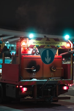 A highway maintenance truck is seen from the back, illuminated at night. The vehicle has a sign with diagonal stripes and the words 'Highway Maintenance' on top. Below the sign, a large arrow points downward. The truck has red and orange lights, as well as reflective materials, indicating it is designed for visibility. Equipment and containers are visible on the back, suggesting the vehicle is equipped for road work.