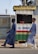 Two individuals are walking past a small kiosk with signage for the National Lottery Authority. The kiosk, decorated with a sign that reads 'Full Payment,' features yellow, green, red, and white colors. The individuals are dressed in matching long blue robes and hats, with one wearing black boots and the other white boots.