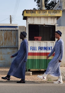 Two individuals are walking past a small kiosk with signage for the National Lottery Authority. The kiosk, decorated with a sign that reads 'Full Payment,' features yellow, green, red, and white colors. The individuals are dressed in matching long blue robes and hats, with one wearing black boots and the other white boots.