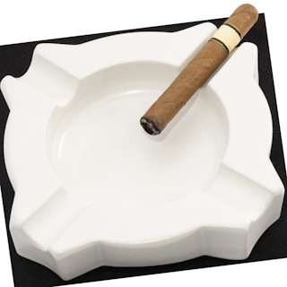 A white ceramic ashtray with an unused cigar resting on its edge. The ashtray is empty and has a smooth, glossy finish. The cigar is light brown with a darkened, slightly burnt tip.