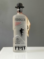 A bottle of Fiddler Vodka is displayed, featuring a frosted glass design with silhouette of a fiddler and dancers. It has a black cap shaped like a bowler hat and a tag attached to the neck. The label includes details of its production in Krakow and the alcohol volume.