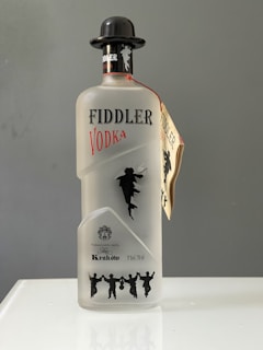 A bottle of Fiddler Vodka is displayed, featuring a frosted glass design with silhouette of a fiddler and dancers. It has a black cap shaped like a bowler hat and a tag attached to the neck. The label includes details of its production in Krakow and the alcohol volume.