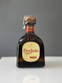 A bottle of Don Julio Reposado tequila with a brown cap and yellow label, placed on a reflective surface against a gray background.