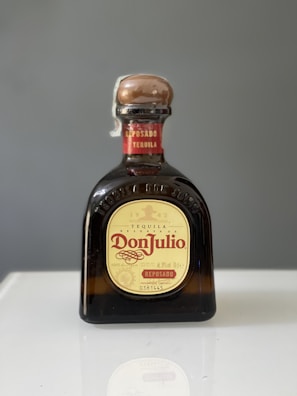 A bottle of Don Julio Reposado tequila with a brown cap and yellow label, placed on a reflective surface against a gray background.