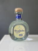 A bottle of Don Julio Blanco tequila with a blue and cream label is centered on a reflective surface. The bottle features a wooden cork and the engraving '1942' above the label.