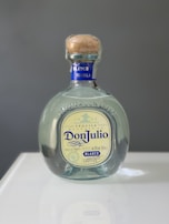A bottle of Don Julio Blanco tequila with a blue and cream label is centered on a reflective surface. The bottle features a wooden cork and the engraving '1942' above the label.