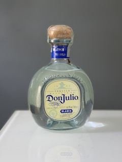 A bottle of Don Julio Blanco tequila with a blue and cream label is centered on a reflective surface. The bottle features a wooden cork and the engraving '1942' above the label.