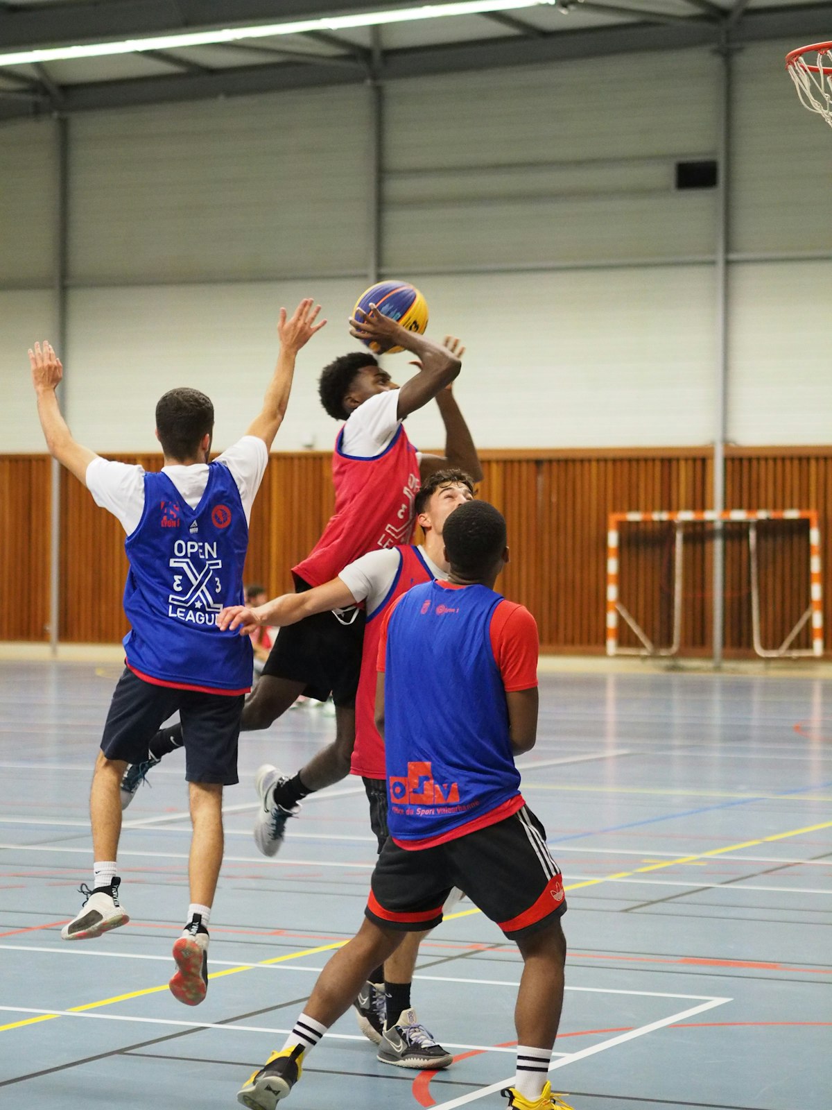 Match de basket 3x3 dans un gymnase éclairé aux néons, un joueur tirant en suspension face à la défense : photo de sport en salle