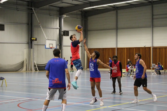 Several people are playing an indoor basketball game on a large court featuring a high ceiling and a scoreboard showing 12:01. A player in a red jersey is leaping to shoot the basketball while another in a blue jersey attempts to block. Three additional players and two people at a table are visible in the background.