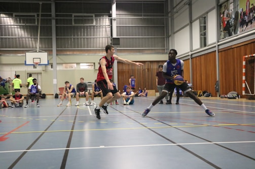 A basketball game is taking place in an indoor court. Two players are in the foreground, one in a red jersey and the other in a blue jersey, engaged in a play. Several other players are seated on the sidelines watching the game. The environment is a gymnasium with a basketball hoop visible on the left and large windows on the upper part of the wall.