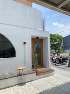 A modern building exterior featuring a white facade with a wooden door and a small rectangular window. A cactus in a pot is placed beside the door on a brown doormat. A simple wooden bench and round table are situated on a pebble and tile pathway. Several motorcycles are parked in the background near the street area.