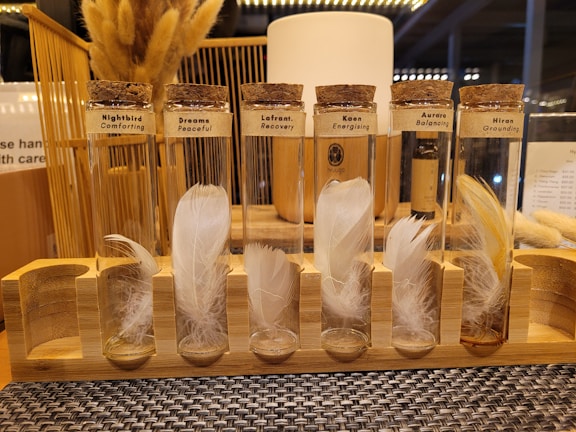 A display of seven test tubes with cork stoppers, each containing delicate white or light-colored feathers. The tubes are labeled with different themes like Comforting, Peaceful, and Energizing. They are arranged on a wooden stand and the setting seems to have a warm, cozy lighting with additional elements like dried plants in the background.