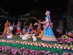 A vibrant and detailed sculpture scene featuring several figures dressed in traditional Indian attire surrounded by nature elements. The figures are depicted in various poses, some holding objects like trays and musical instruments. Peacocks and swans add to the serene and culturally rich environment.