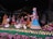 A vibrant and detailed sculpture scene featuring several figures dressed in traditional Indian attire surrounded by nature elements. The figures are depicted in various poses, some holding objects like trays and musical instruments. Peacocks and swans add to the serene and culturally rich environment.