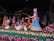 A vibrant and detailed sculpture scene featuring several figures dressed in traditional Indian attire surrounded by nature elements. The figures are depicted in various poses, some holding objects like trays and musical instruments. Peacocks and swans add to the serene and culturally rich environment.
