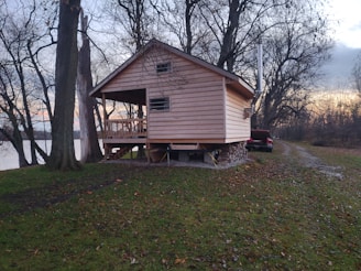 A wooden cabin is elevated on stilts, surrounded by bare trees in a natural setting. There is a grassy area leading to the cabin, with a small road on the right side where a red pickup truck is parked. The backdrop features a body of water, and the sky appears to be at dusk, showcasing a gradient of blue and orange hues.