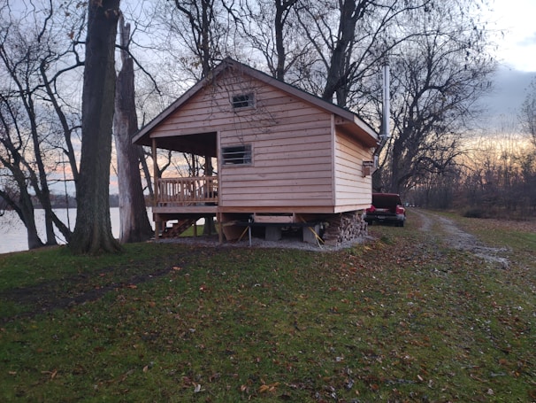 A wooden cabin is elevated on stilts, surrounded by bare trees in a natural setting. There is a grassy area leading to the cabin, with a small road on the right side where a red pickup truck is parked. The backdrop features a body of water, and the sky appears to be at dusk, showcasing a gradient of blue and orange hues.